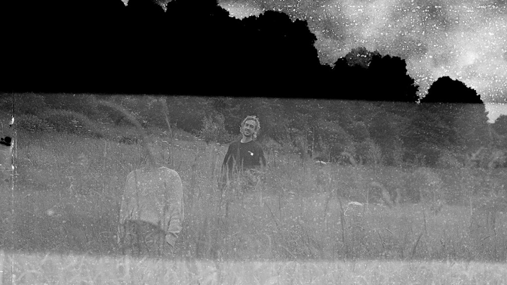 a monotone analogue photograph showing a headless figure in the foreground, a male in the background and a treeline in the background.